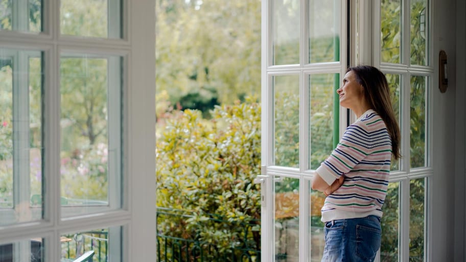 A woman looking out a white terrace door onto a green bush